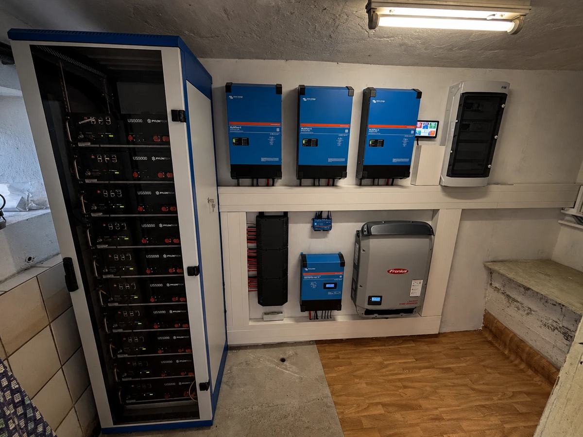 Technikraum der Anlage: links ein offener blau-weißer Batterieschrank mit zehn übereinander eingeschobenen Pylontech US5000, rechts an der Wand drei blaue Victron MultiPlus-II nebeneinander, daneben ein GX-Touch-Display und die AC-Verteilung. Darunter die schwarze PowerLink-Verteilung mit den roten und schwarzen DC-Leitungen, ein SmartSolar-Laderegler und ein Fronius-Wechselrichter.